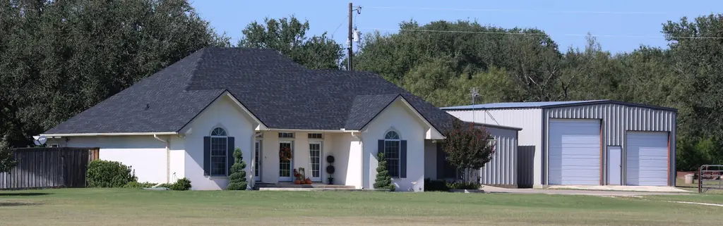 White house with a dark hip shingle roof next to a metal shop building, an Excell Roofing job.
